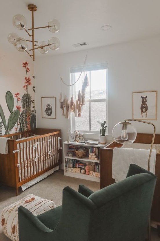 a desert themed twin nursery with stained cribs, cactus on the wall and dresser, a catchy chandelier and a green chair
