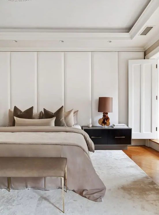 a fabulous contemporary bedroom with a headboard clad with upholstered panels, a bed with neutral earthy bedding and a matching bench, a large nightstand with a lamp