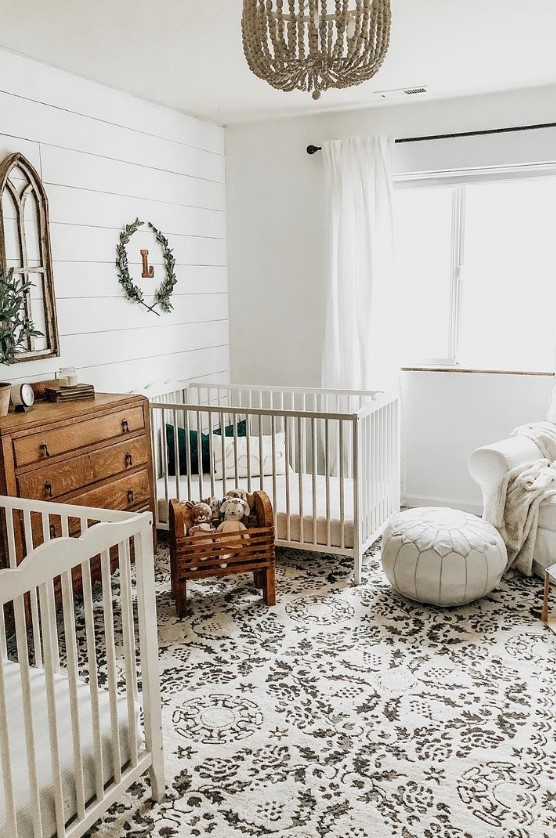 a farmhouse twin nursery with white cribs, a stained dresser, a wooden bead chandelier, some fake greenery and a vintage window frame