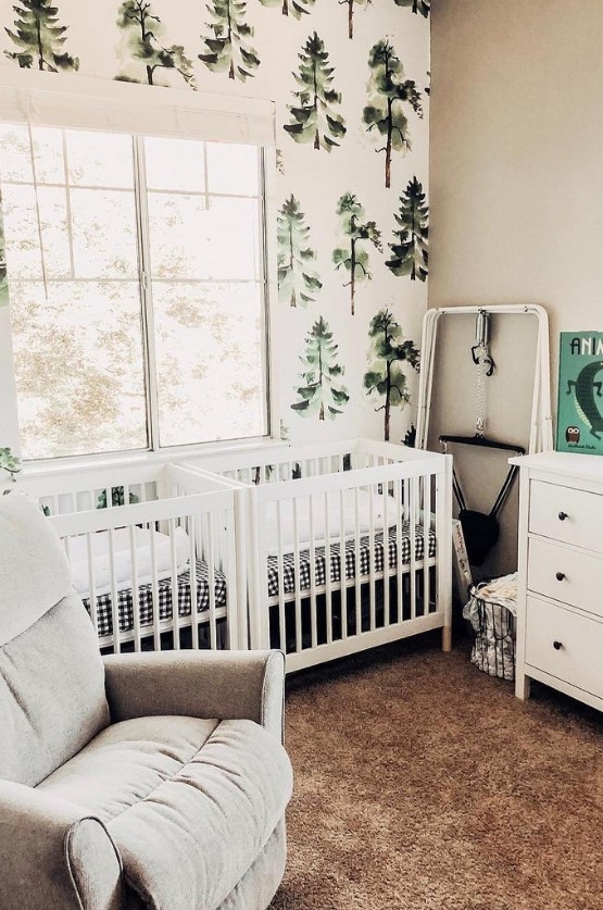 a forest-themed twin nursery with white cribs, tree-printed wallpaper, white and grey furniture and a pretty soft rug