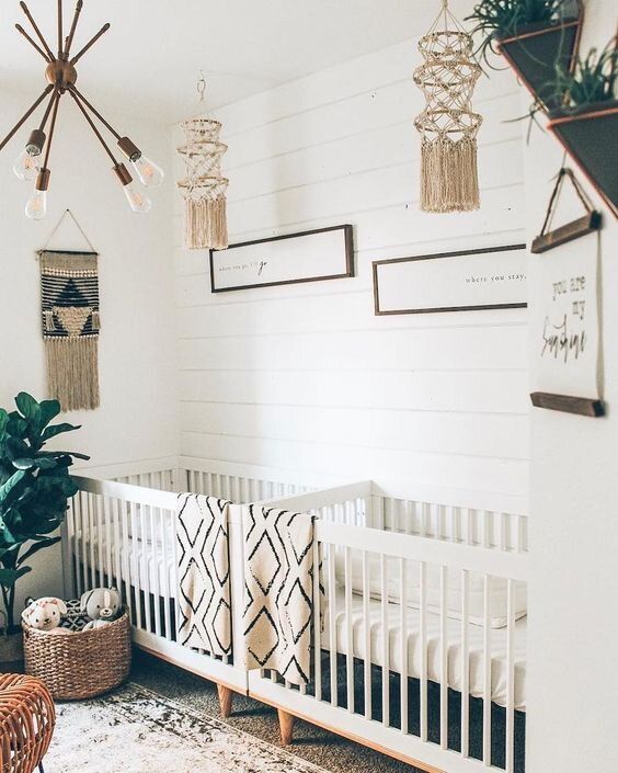a gender neutral boho twin nursery with shiplap walls, white cribs, woven mobiles, a modern chandelier, potted plants and a basket for toys