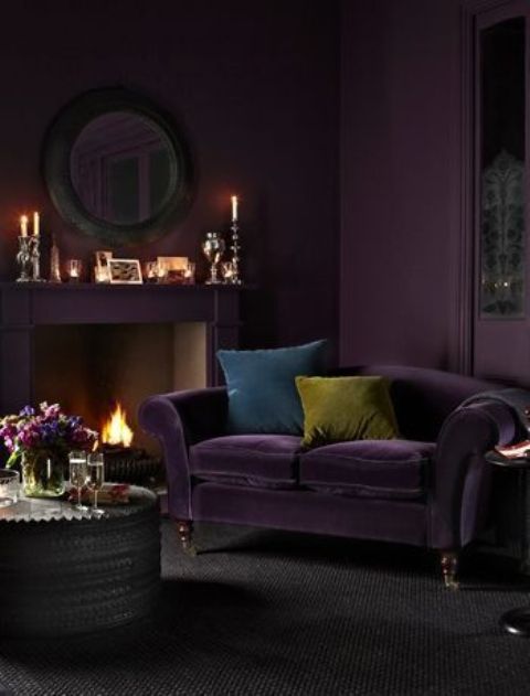 a gorgeous aubergine living room with a matching fireplace, a round mirror, candles in chic candleholders, a deep purple loveseat and a side table