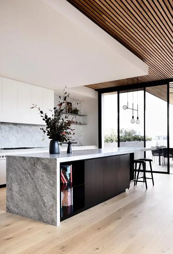 a gorgeous contemporary ktichen with sleek white cabinets, a grey stone backsplash and countertops, a black kitchen island and a layered ceiling