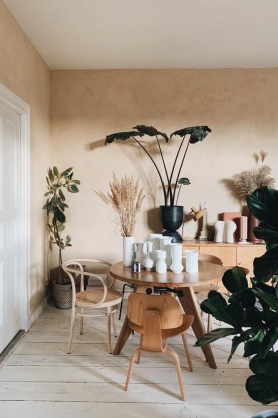a gorgeous dining space with tan limewashed walls, a light-stained vanity, a wooden table and mismatching chairs, lots of plants