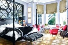 a gorgeous maximalist bedroom with lilac walls, a black bed and an ottoman, red chairs and a grey windowsill bench, a crystal chandelier