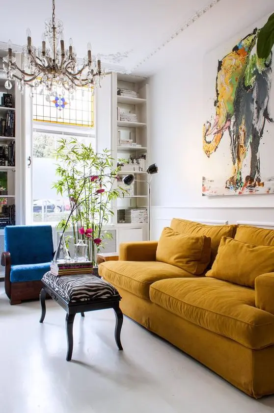 a gorgeous maximalist living room with a mustard sofa, a blue chair, potted plants, a refined chandelier and a bold artwork