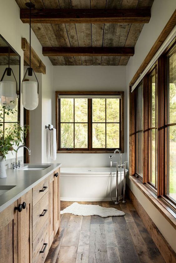 a gorgeous modern rustic bathroom with a wooden ceiling and beams, a wooden floor and a vanity, large windows and a bathtub