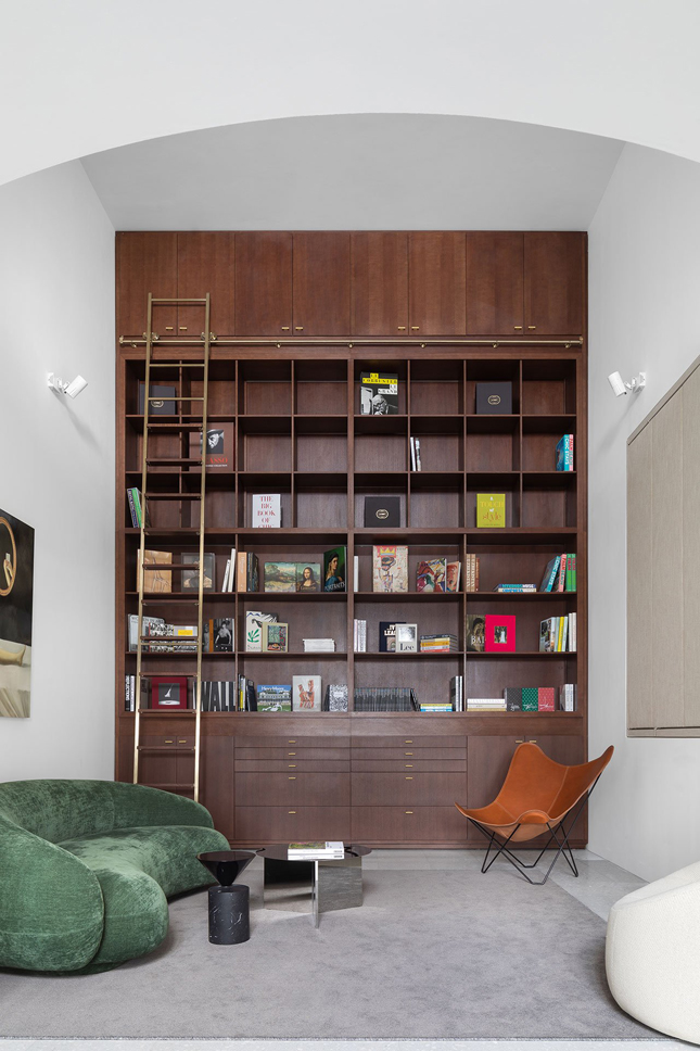 a home library with a large bookcase and a ladder, a green curved low sofa, a creamy chair and a leather one, side tables