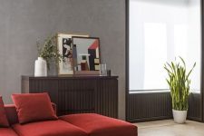 a laconic modern living room with grey walls, a deep red sectional, a wooden storage unit and a low round table
