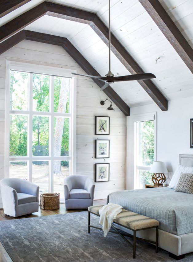 a large airy farmhouse bedroom done with white walls, dark stained ceiling beams and comfy furniture