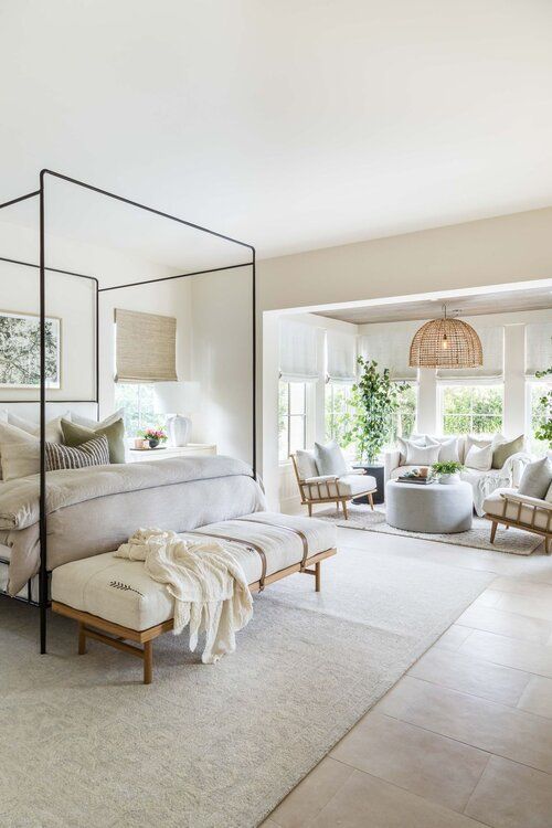 a large neutral bedroom with a frame bed with neutral bedding, a creamy bench with a blanket and a large seating zone by the window