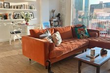 a light-filled living room with open shelves, a desk and a chair, an orange sectional sofa, a coffee table and colorful pillows