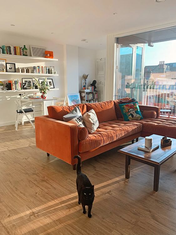 a light filled living room with open shelves, a desk and a chair, an orange sectional sofa, a coffee table and colorful pillows