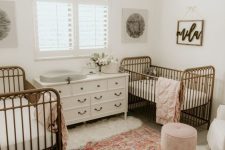 a little ladies’ twin nursery with vintage metal cribs, a large dresser as a changing table, a printed rug, a pink pouf and some pretty art