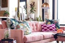 a lively living room with a pink sofa, floral pillows and a rug, stacks of books, a gold floor lamp, greenery and candles plus lots of natural light