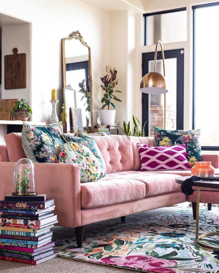a lively living room with a pink sofa, floral pillows and a rug, stacks of books, a gold floor lamp, greenery and candles plus lots of natural light