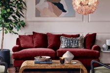 a lovely blush living room with paneled walls, a deep red sofa, a wooden coffee table, green chairs, a printed rug, an abstract artwork and a potted tree