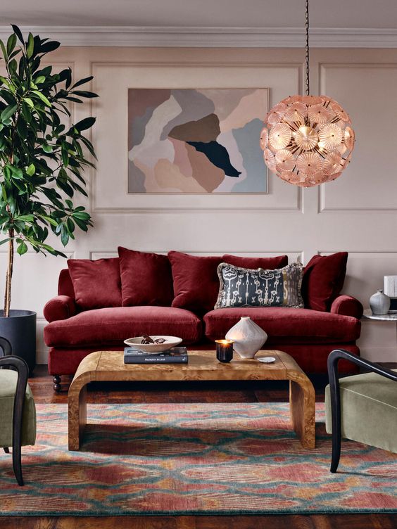a lovely blush living room with paneled walls, a deep red sofa, a wooden coffee table, green chairs, a printed rug, an abstract artwork and a potted tree