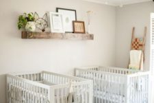 a lovely farmhouse twin nursery with grey walls, white cribs, a stained shelf with artworks and potted greenery plus a ladder