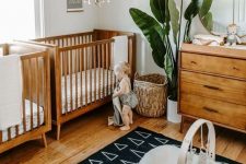 a lovely gender neutral boho twin nursery with stained cribs and a dresser, a woven basket, a potted plant, a black and white geo printed rug and a round mirror