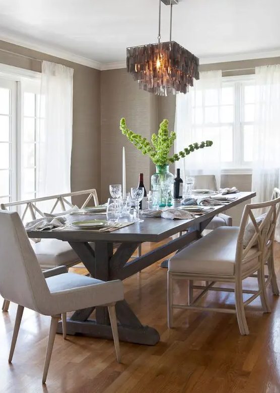 a lovely neutral dining room with farmhouse touches, with greige grasscloth wallpaper, a trestle table and neutral chairs and a tassel chandelier