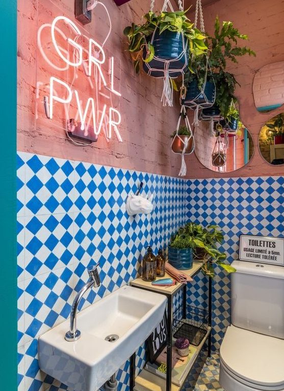 a maximalist bathroom with pink brick walls, blue and white checked tiles, a console table, potted greenery, a neon sign and mirrors