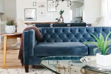 a mid-century modern living room with a blue velvet sofa, a glass coffee table, a bold printed rug and potted greenery
