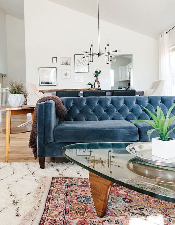 a mid-century modern living room with a blue velvet sofa, a glass coffee table, a bold printed rug and potted greenery