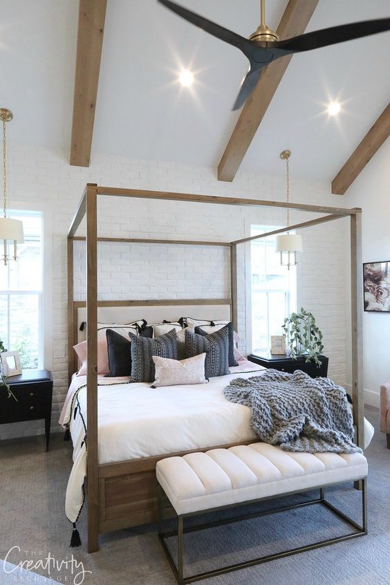 a modern farmhouse bedroom with a faux white brick wall, a frame bed with neutral bedding, a white bench and wodoen beams on the ceiling