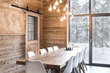 a modern rustic dining room with the ceiling, walls and a floor clad with wood, a wooden dining table, white Eames chairs and a bulb chandelier