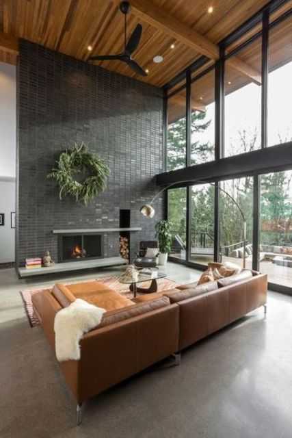 a modern rustic living room with a glazed wall, a fireplace wall clad with black tiles, an amber leather corner sofa, a glass table and a floor lamp