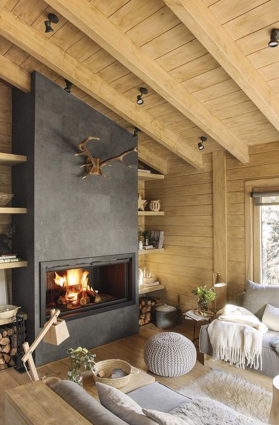 a modern rustic living room with wooden walls and a ceiling, a fireplace, firewood stored by it and antlers