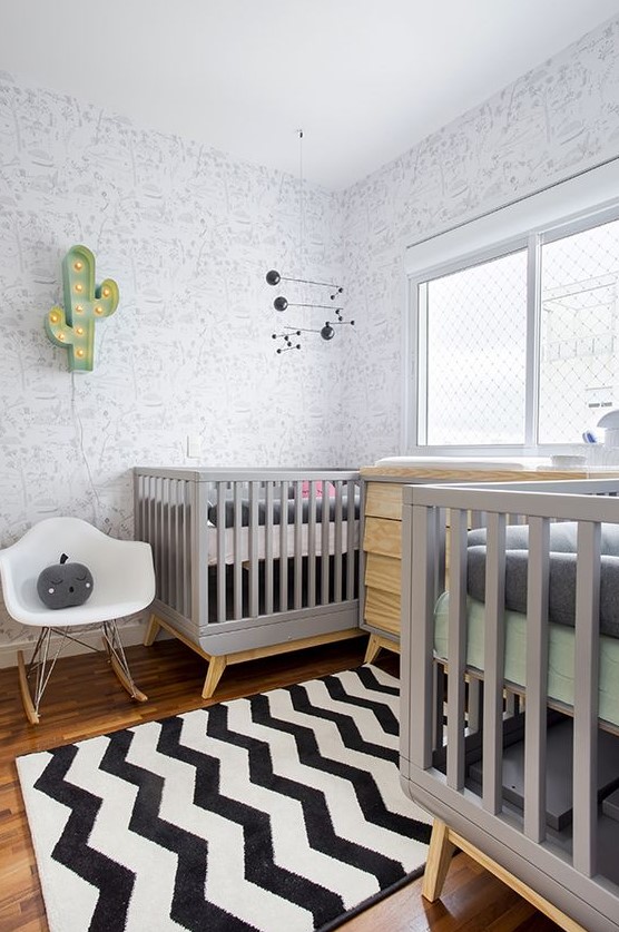 a modern twin nursery with printed walls, grey cribs, a stained dresser, a pritned rug, a white chair and a cactus lamp