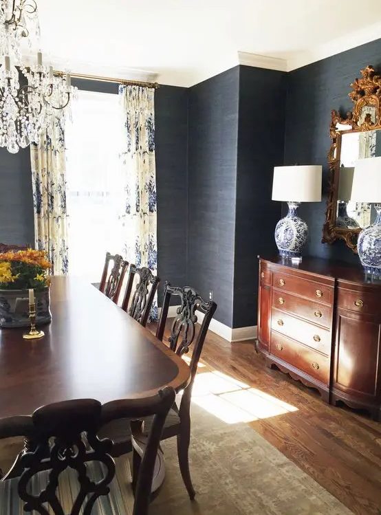 a moody and refined vintage dining room with black grasscloth wallpaper, dark-stained vintage furniture, a mirror in an ornated frame and a crystal chandelier