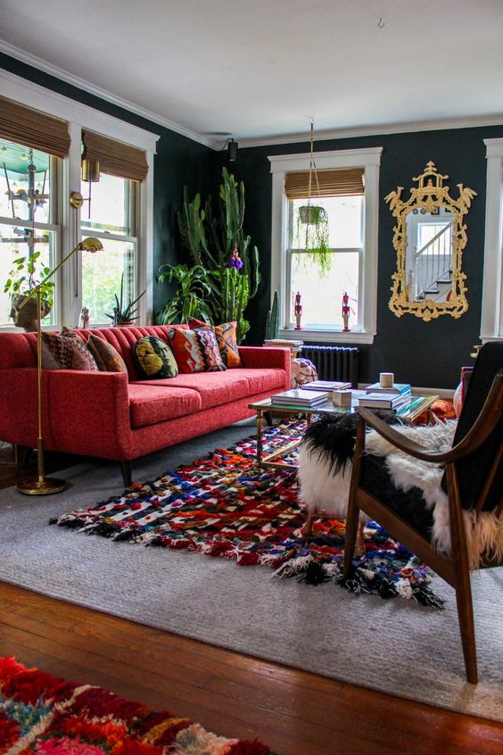 a moody black living room with a red sofa, a glass table, a black chair, a colorful rug, potted cacti and plants, a mirror in a gold frame