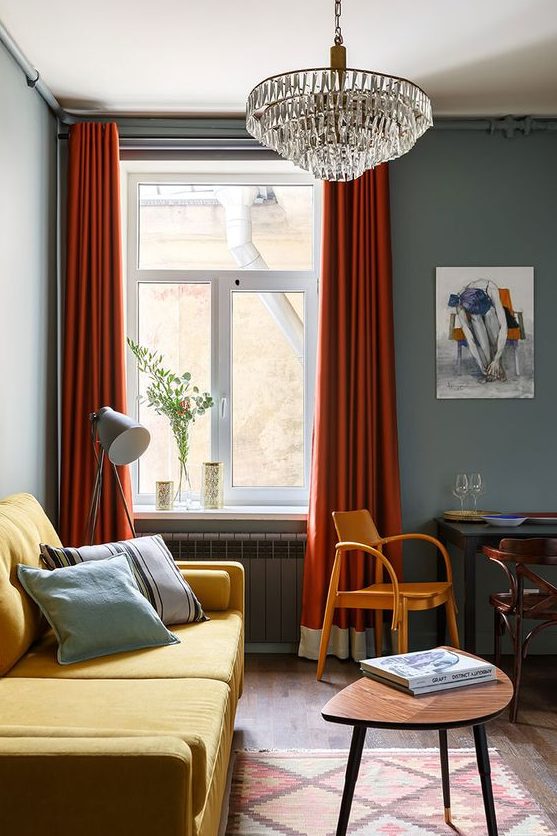 a moody living room with grey walls, a yellow sofa, rust-colored curtains, a crystal chandelier and vintage chairs and tables