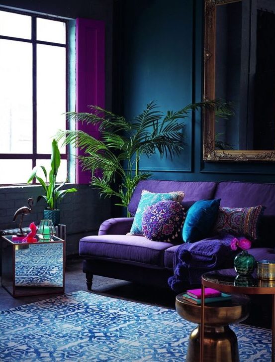 a moody living room with navy paneled walls, a purple sofa, metallic tables and a mirror one, a blue rug and hot pink curtains