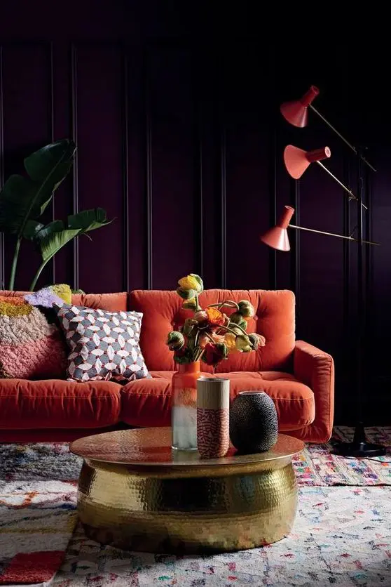 a moody living room with purple paneled walls, an orange sofa, orange floor lamps, a gold hammered table and statement plants