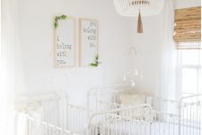 a neutral and gender neutral twing nursery with white vintage cribs, artwork, a wooden bead chandelier and some rustic blinds