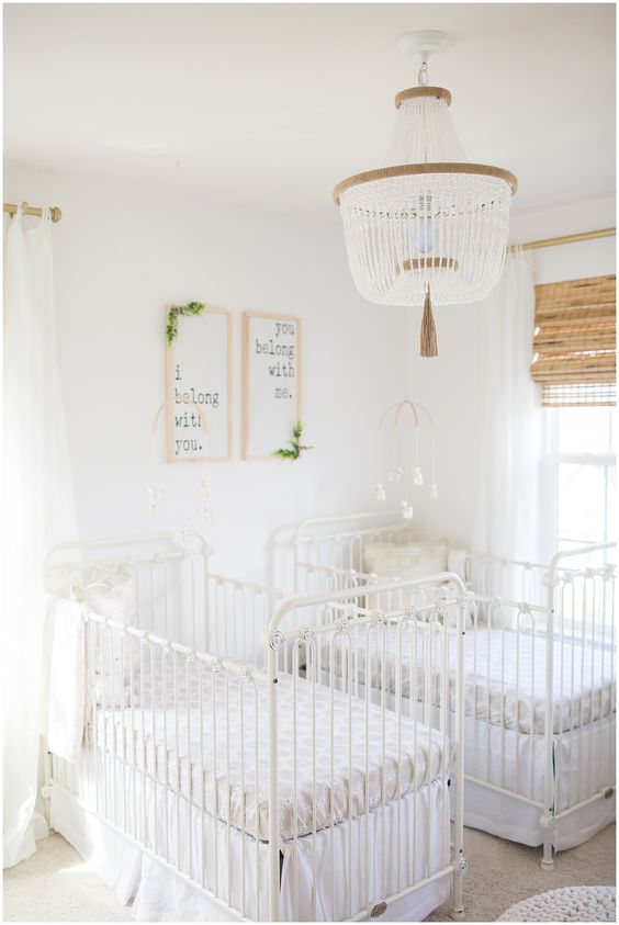 a neutral and gender neutral twing nursery with white vintage cribs, artwork, a wooden bead chandelier and some rustic blinds