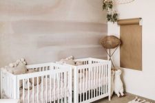 a neutral twin nursery with a statement wall, a couple of white cribs, a woven lampshade, some layered rugs and a white chair with a footrest