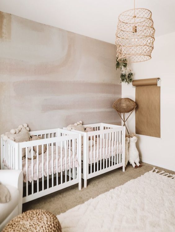 a neutral twin nursery with a statement wall, a couple of white cribs, a woven lampshade, some layered rugs and a white chair with a footrest
