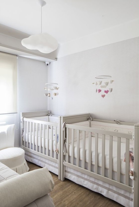 a neutral twin nursery with a wallpaper wall, grey cribs and a creamy sofa plus a creamy ottoman is very cool