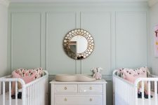 a pastel twin nursery with green paneled walls, white cribs, pink bedding, a white dresser as a changing table, a chic mirror, a floral printed rug