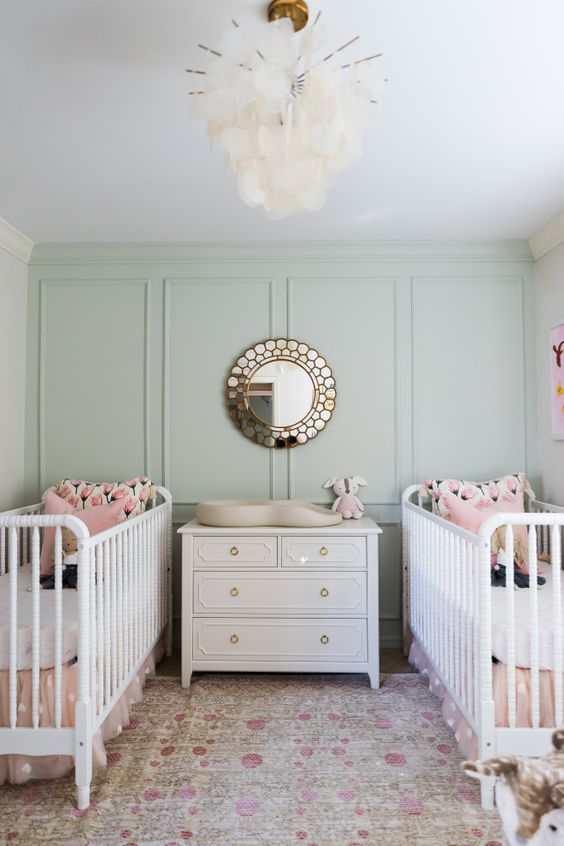 a pastel twin nursery with green paneled walls, white cribs, pink bedding, a white dresser as a changing table, a chic mirror, a floral printed rug