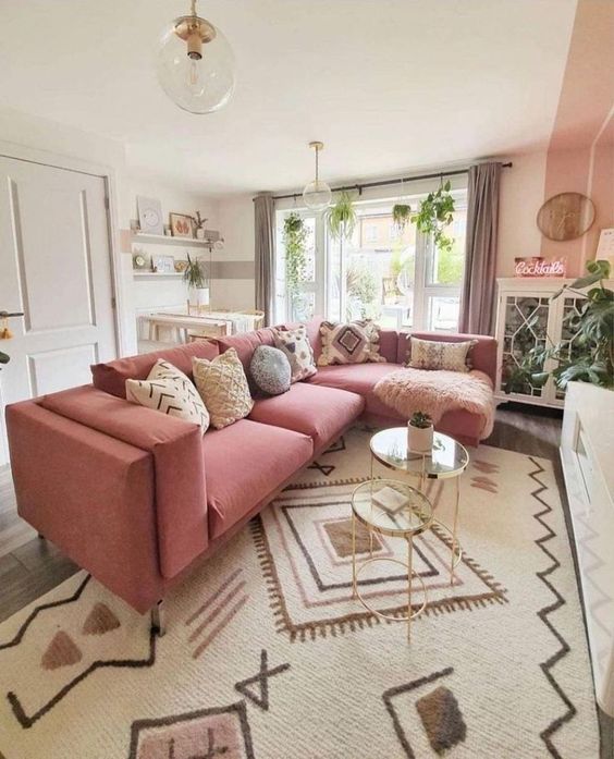a pretty boho living room done in neutrals, with a large pink sectional in front of the fireplace, glass tables, greenery and a working space by the window