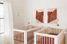a pretty boho mid-century modern nursery with white cribs, terracotta bedding and macrame, a rattan lamp and a printed rug