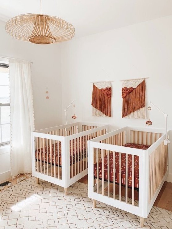 a pretty boho mid-century modern nursery with white cribs, terracotta bedding and macrame, a rattan lamp and a printed rug