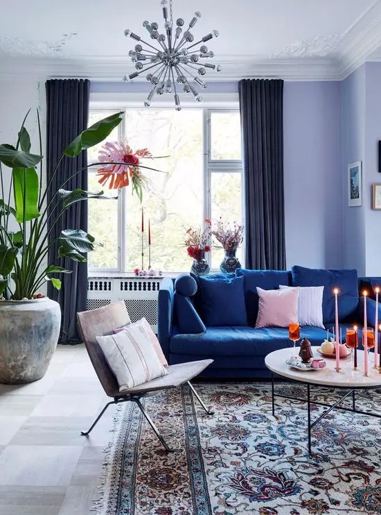 a pretty living room done with a cold color scheme, with lilac walls, a navy sofa, a round table, a lovely chair, some greenery and a sunburst chandelier