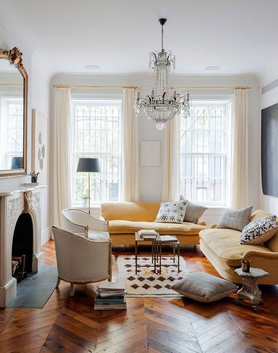 a refined Parisian living room with a non-working fireplace, a yellow sectional, a refined crystal chandelier, creamy chairs and a large mirror over the fireplace